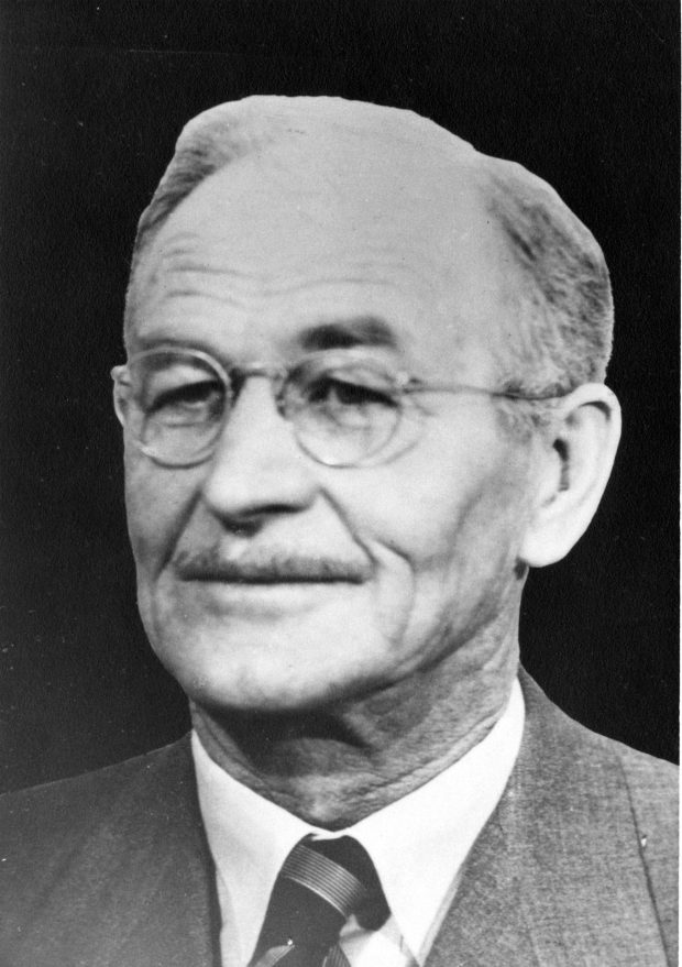 Black and white photograph of a man in a suit and tie wearing glasses and a thin moustache.