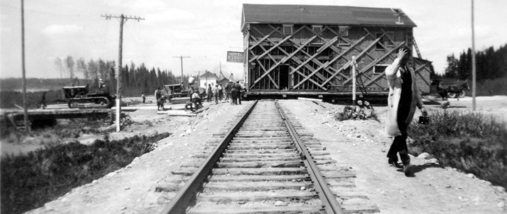 Black and white photograph of a two-storey building being moved across a railway. Hotel St-Louis’ sign is visible. A dozen curious onlookers watch two tractors haul the building.