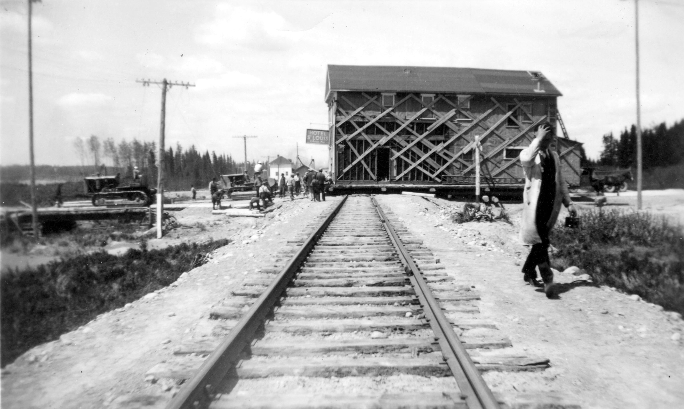 Black and white photograph of a two-storey building being moved across a railway. Hotel St-Louis’ sign is visible. A dozen curious onlookers watch two tractors haul the building.