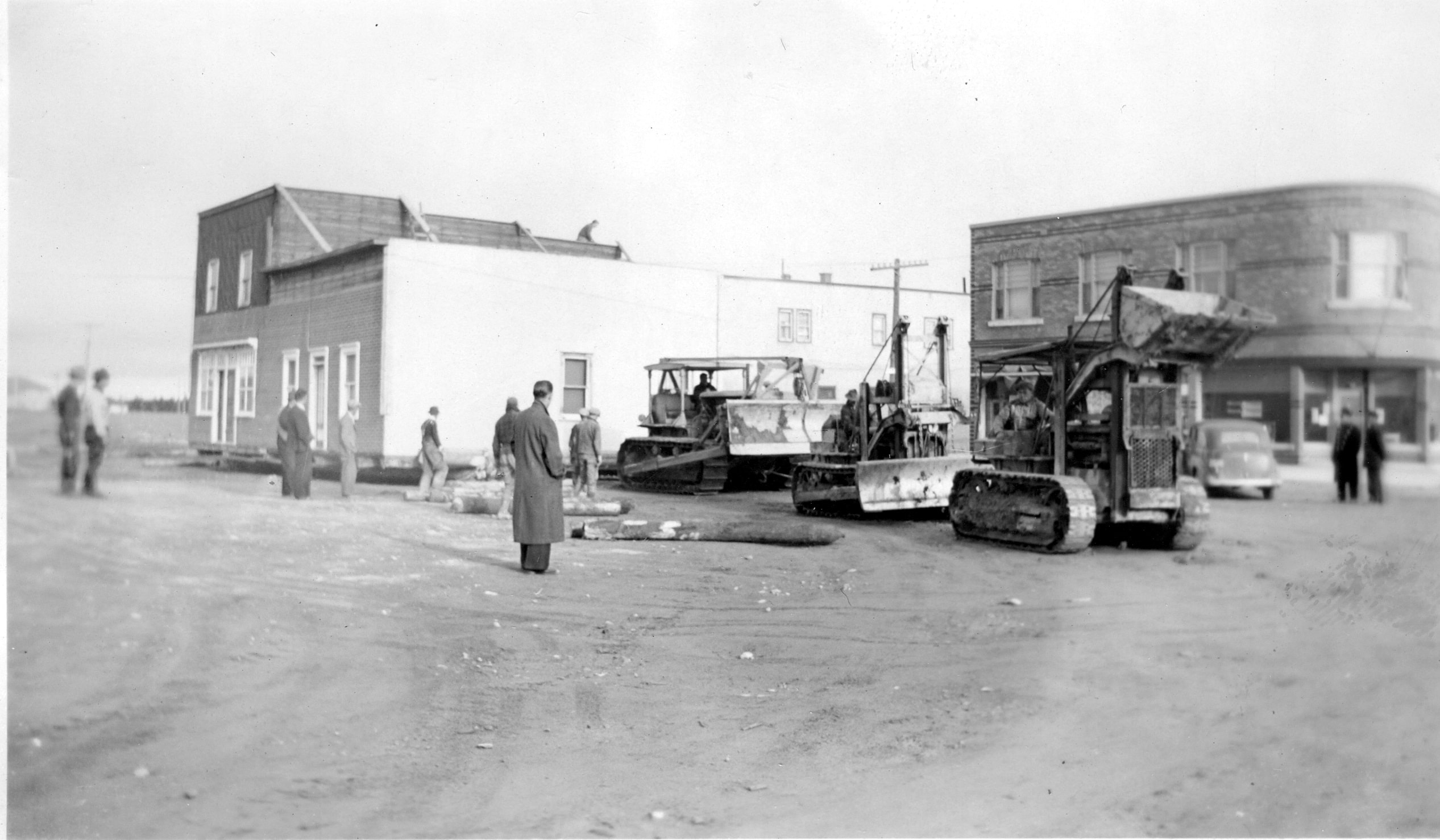 Black and white photograph of a two-storey building being pulled by three tractors. The logs used to roll the building are laid down in front. A person is on the roof. Several curious onlookers attend the scene.