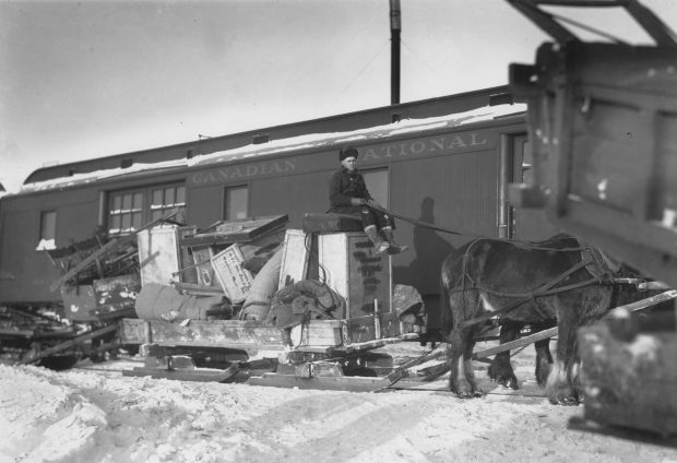 Black and white photograph of a cart, filled to the brim with furniture and boxes, being pulled by two horses driven by a man sitting on the goods. Given the snow, the cart is on skis and the man is dressed warmly. A wagon bearing the inscription Canadian National” is in the background.