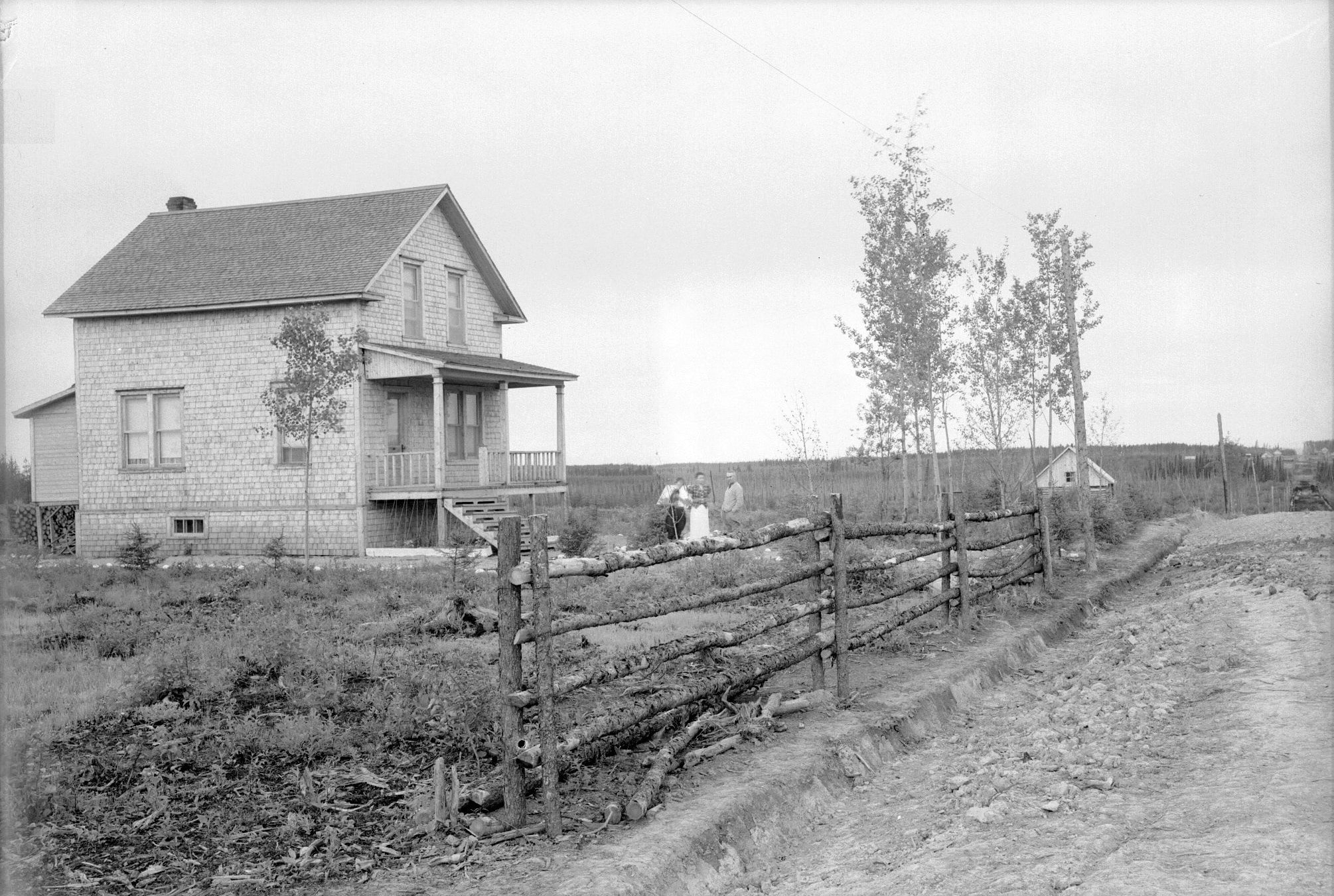 Black and white photograph of a two-storey house with three people posing in front of it. In the foreground, a fence. In the background, a small cluster of houses.
