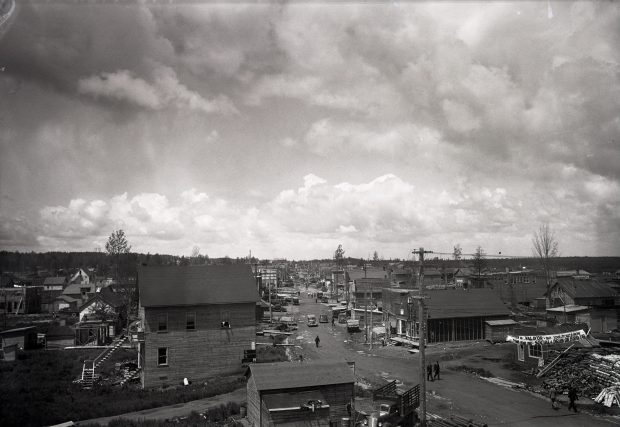Black and white photograph of a street with rudimentary buildings on either side. Cars and pedestrians are out and about in the centre of the unpaved roadway, which is dotted with holes full of water.