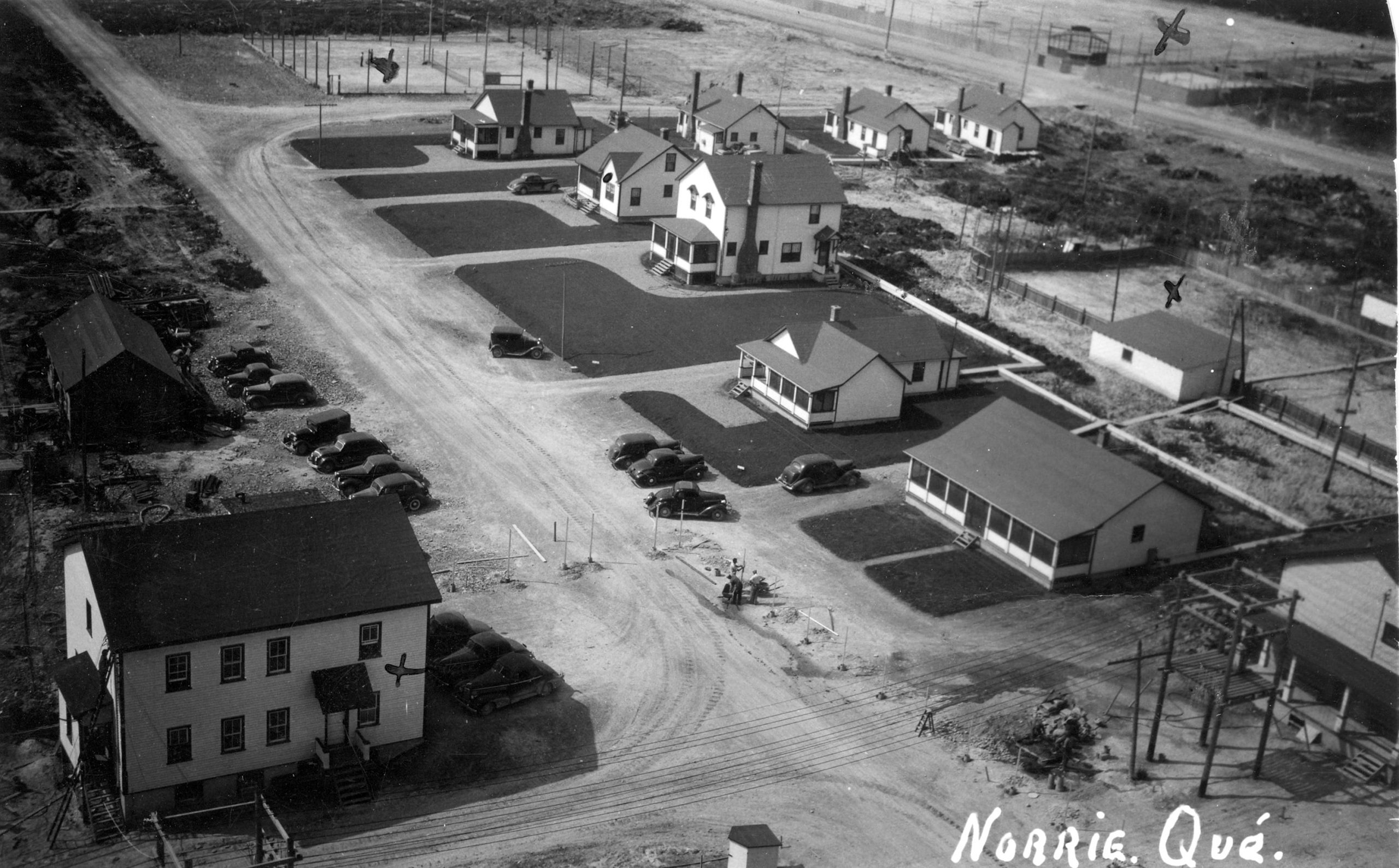 Black and white photograph of a hamlet consisting of several modern buildings. Several cars are parked in front of well-manicured lawns. Several sports fields (baseball, hockey, and tennis) are also visible.