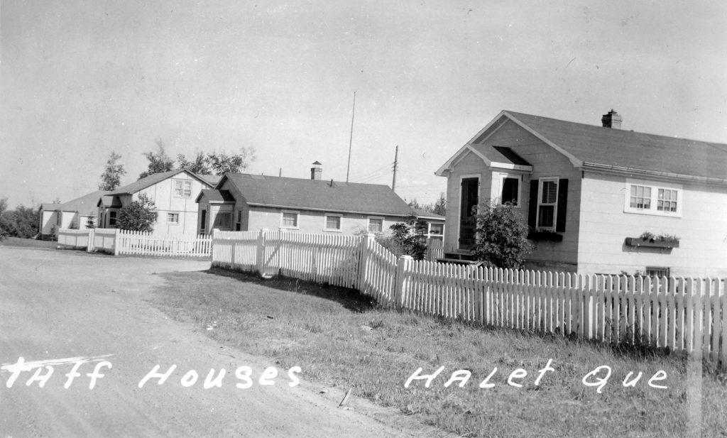 Photographie en noir et blanc de plusieurs résidences construites de planches qui sont en-tourées d’une clôture blanche. Au bas, l’inscription « Staff Houses, Halet Que ».