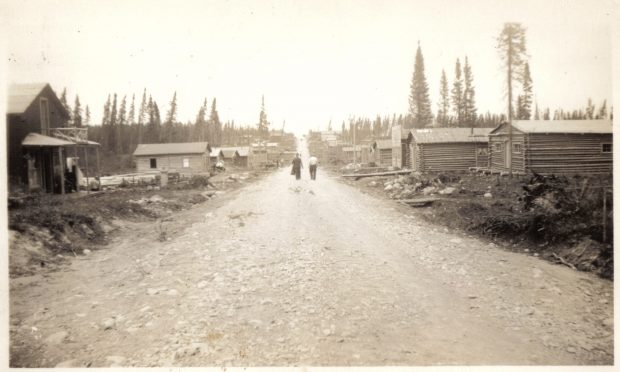 Sepia photograph of a gravel road lined with log cabins. In the centre, two men are walking towards a hill. Several conifers are visible in the background.