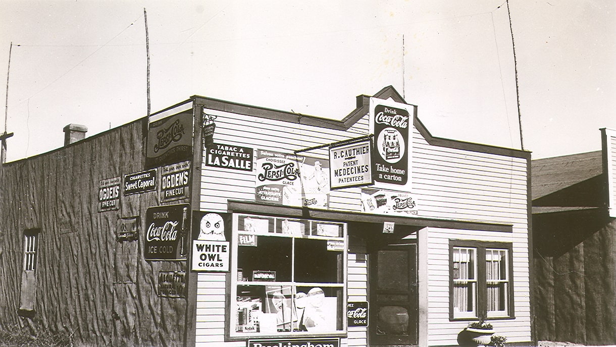 Black and white photograph of a building with a plank façade and tar paper side. A dozen advertisements are displayed, notably for Coca-Cola and various brands of cigarettes (Sweet Caporal and Lasalle).