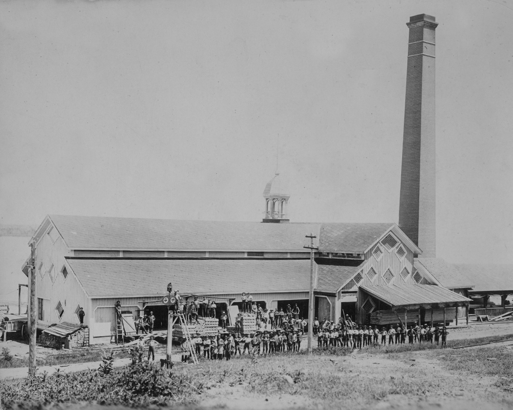 Mill employees pose for a group photo in front of lumber mill located beside a river.