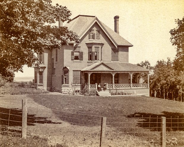 A young couple sits on the steps of a large, brick house with a wrap- around porch.
