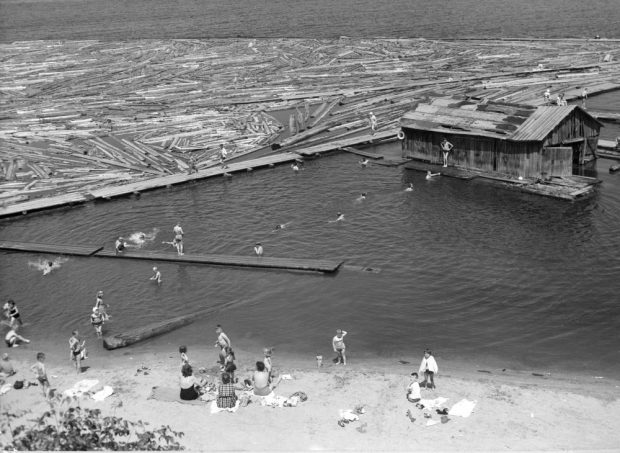 A group of women sit on a sandy beach watching children play and swim to a boat house is a short distance from the shoreline.