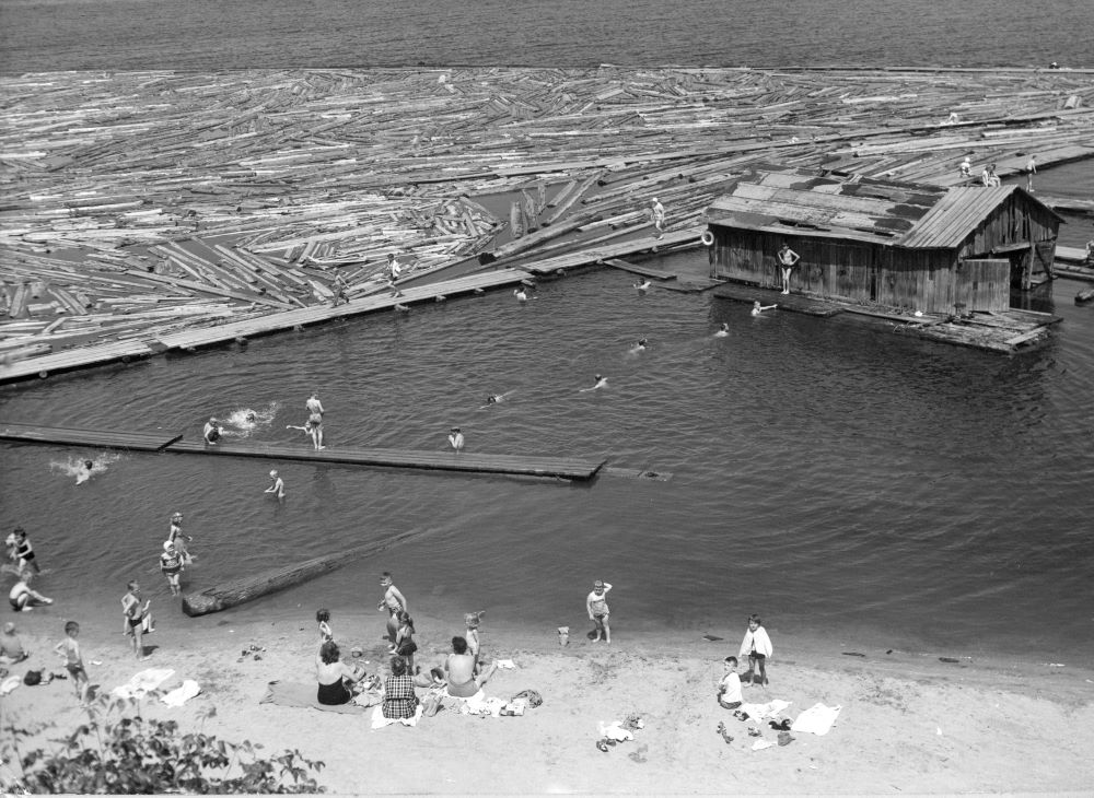 A group of women sit on a sandy beach watching children play and swim to a boat house is a short distance from the shoreline.