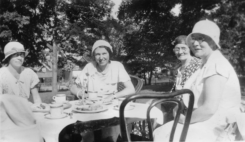 Four ladies wearing hats pose for the camera as they sit outdoors at a table set for a tea party.