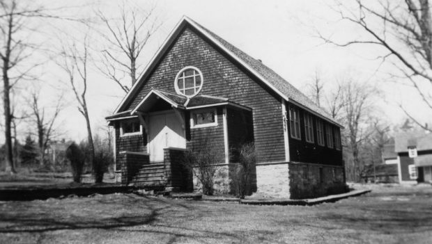 A peaked roof covers the entrance to a small church covered in cedar shingles.