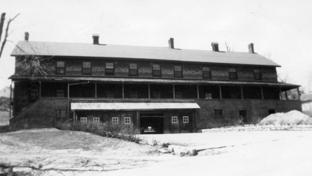 A long building covered in cedar shingles features a porch running the entire length and a garage below where a car is parked.