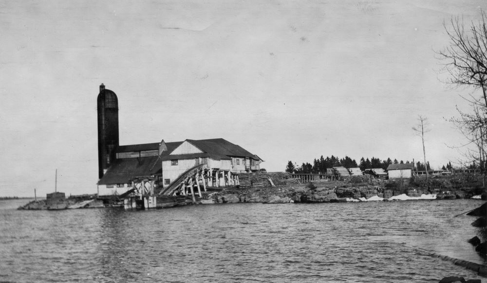 A mill on a rocky shoreline is surrounded by piles of cedar shingles and cut railway ties.