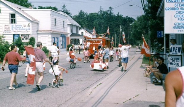 A decorated fire truck leads a parade of children riding bicycles and adults carrying Canadian flags.