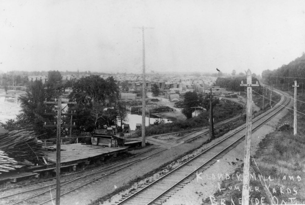 Black and white postcard showing railway tracks running beside a bay with piles of lumber in the distance.