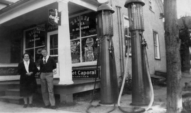 A woman and a young man stand next to old fashioned fuel pumps outside a general store.