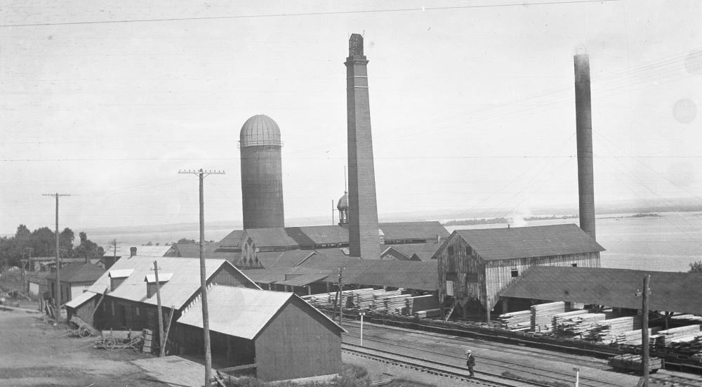 A man stands on a railway track that runs between buildings at a large saw mill site.