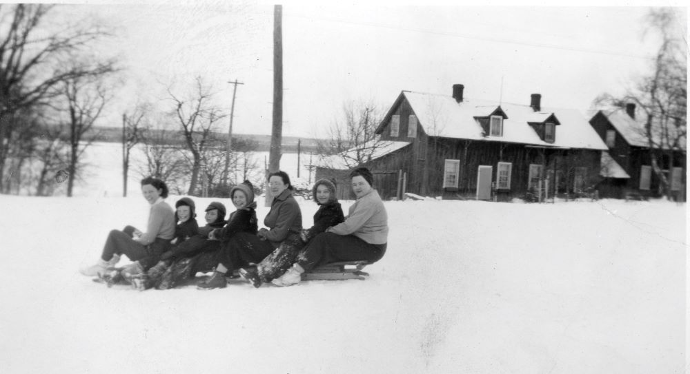 Smiling women and children sit on a long toboggan at the top of a hill.