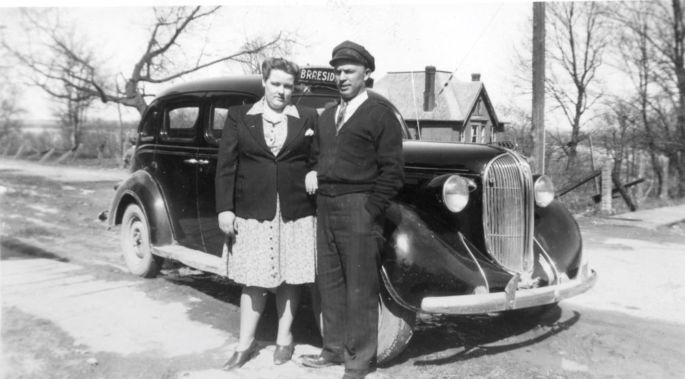 A couple stand on the sidewalk beside a Braeside Taxi car.