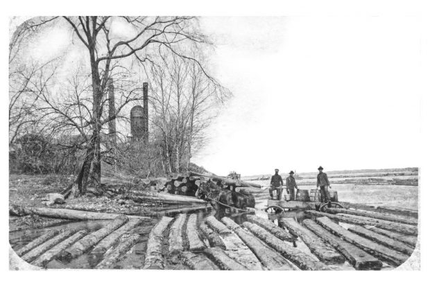 A team of horses wait in the shallow water as three men fill water barrels onto a cart.