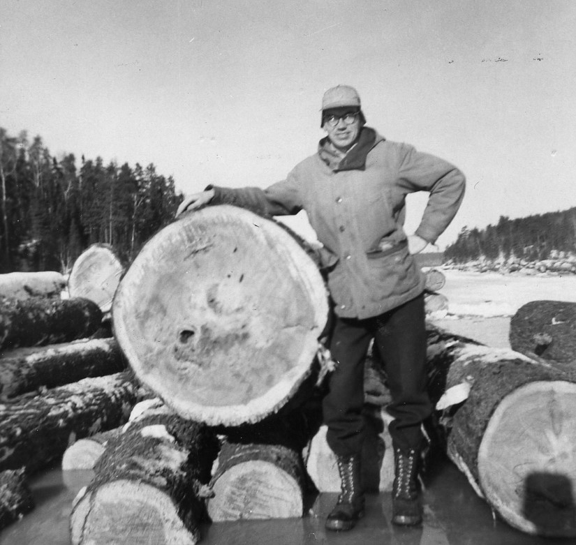 A forester stands on the ice with one hand on his hip and the other draped over a large log.