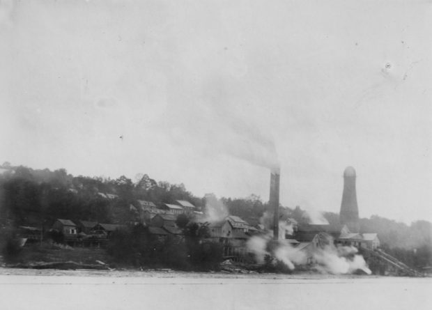 Several buildings surround a mill site on the side of a hill. Steam and smoke from the lake blow off the lake.