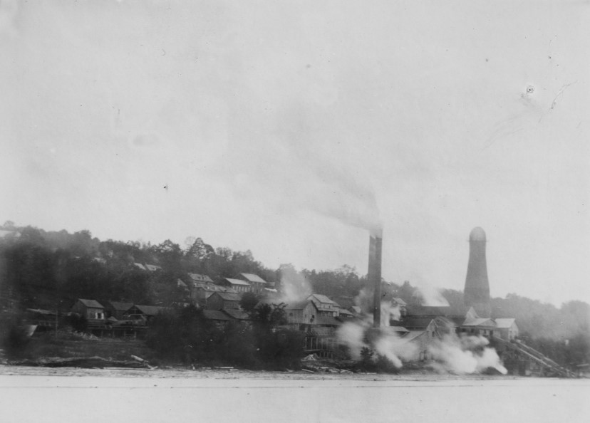 Several buildings surround a mill site on the side of a hill. Steam and smoke from the lake blow off the lake.