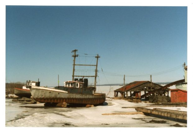 Two old tug boats rest on wooden logs beside a dilapidated wooden shed at the shoreline in winter.