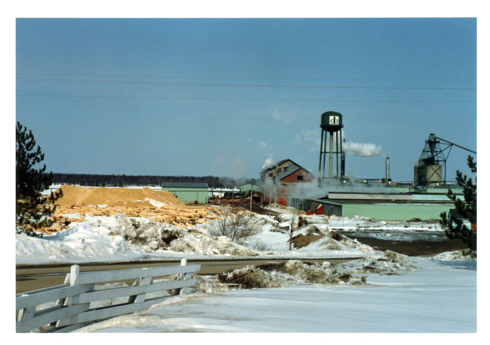 A brick saw mill building is surrounded by heaps of sawdust with a water tower and refuse burner in winter.
