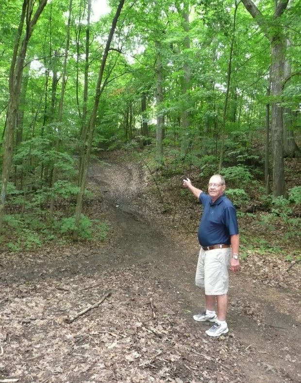 A man wearing shorts points into a wooded area.