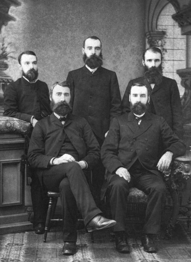 A studio portrait shows three gentlemen standing behind two others who are seated.