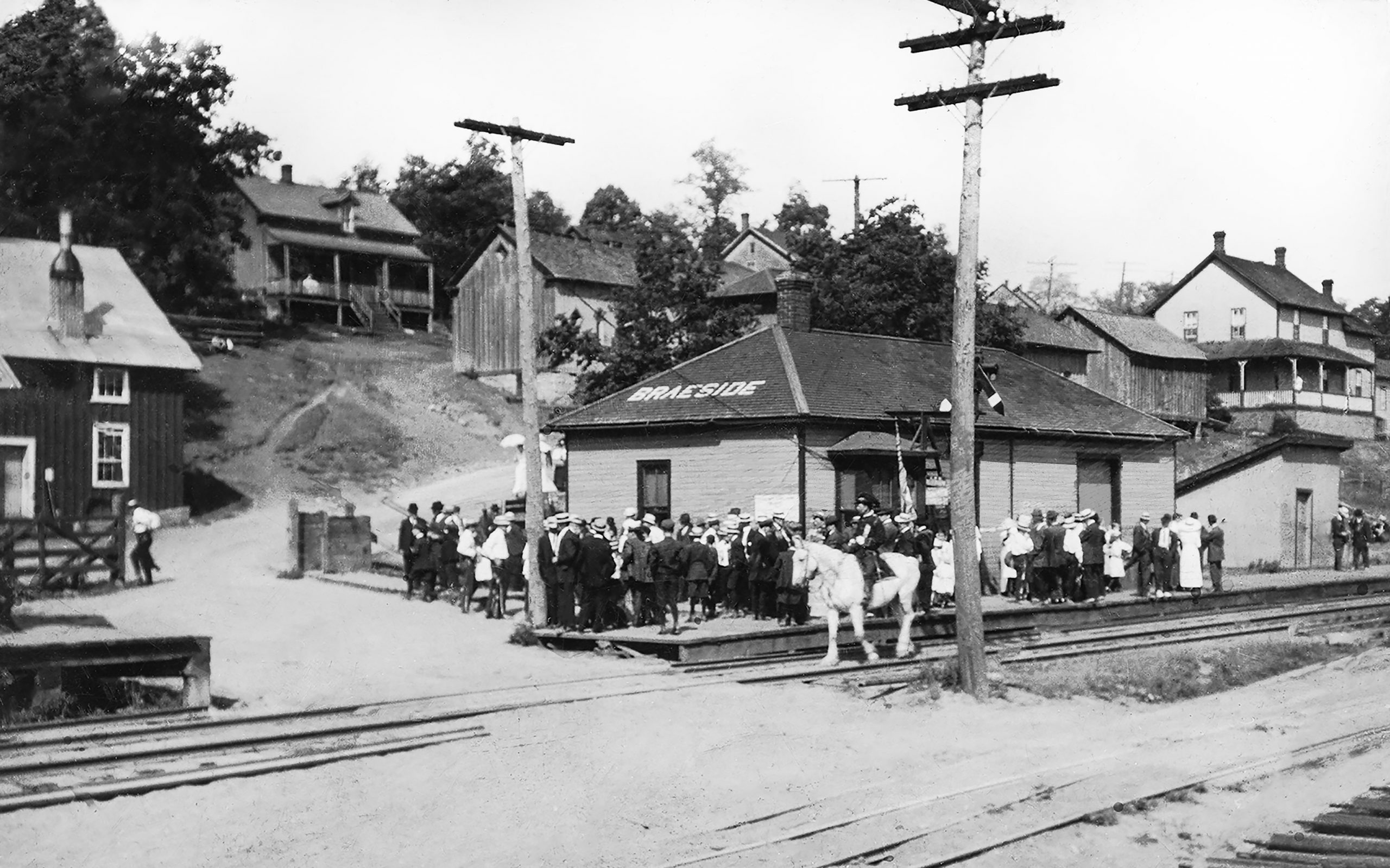 A crowd at the Braeside Train Station watches an Orangeman’s Parade led by a man on a white horse.