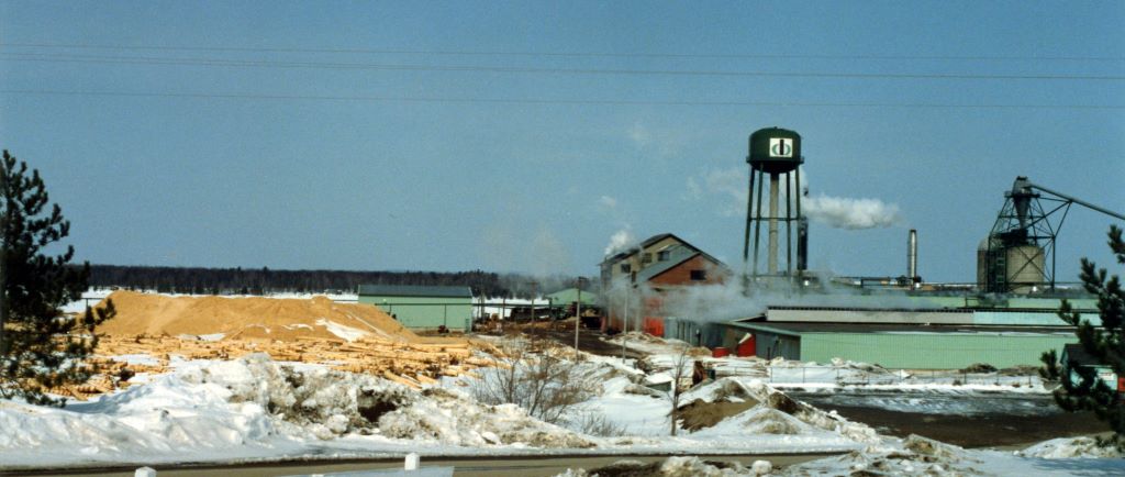 A brick saw mill building is surrounded by heaps of sawdust with a water tower and refuse burner in winter.