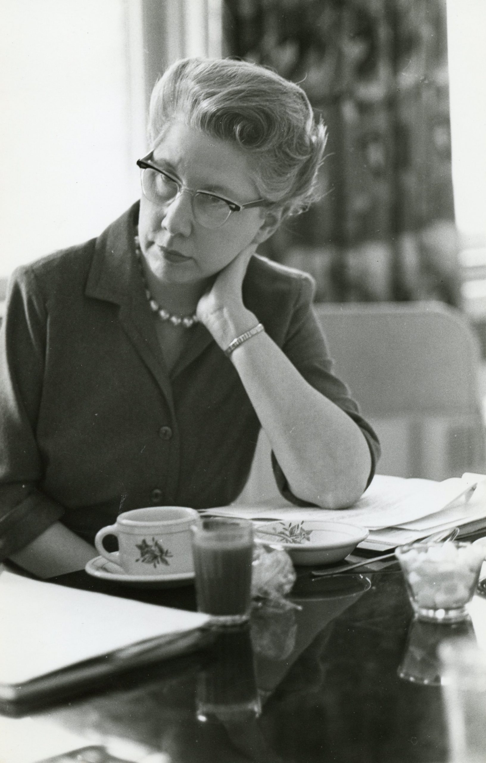 A black and white photo of a woman in a blazer sitting at a table. Before her are papers and the remnants of tea. Her head is propped up by her arm as she listens to something not pictured.