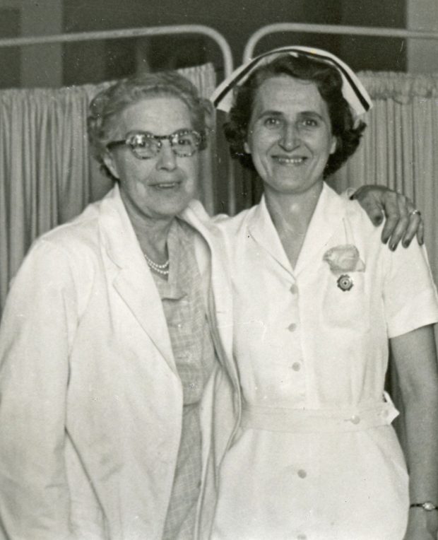 A black and white photo of two women. One wears a lab-coat. She has an arm over the shoulders of a woman in a nurses' uniform.