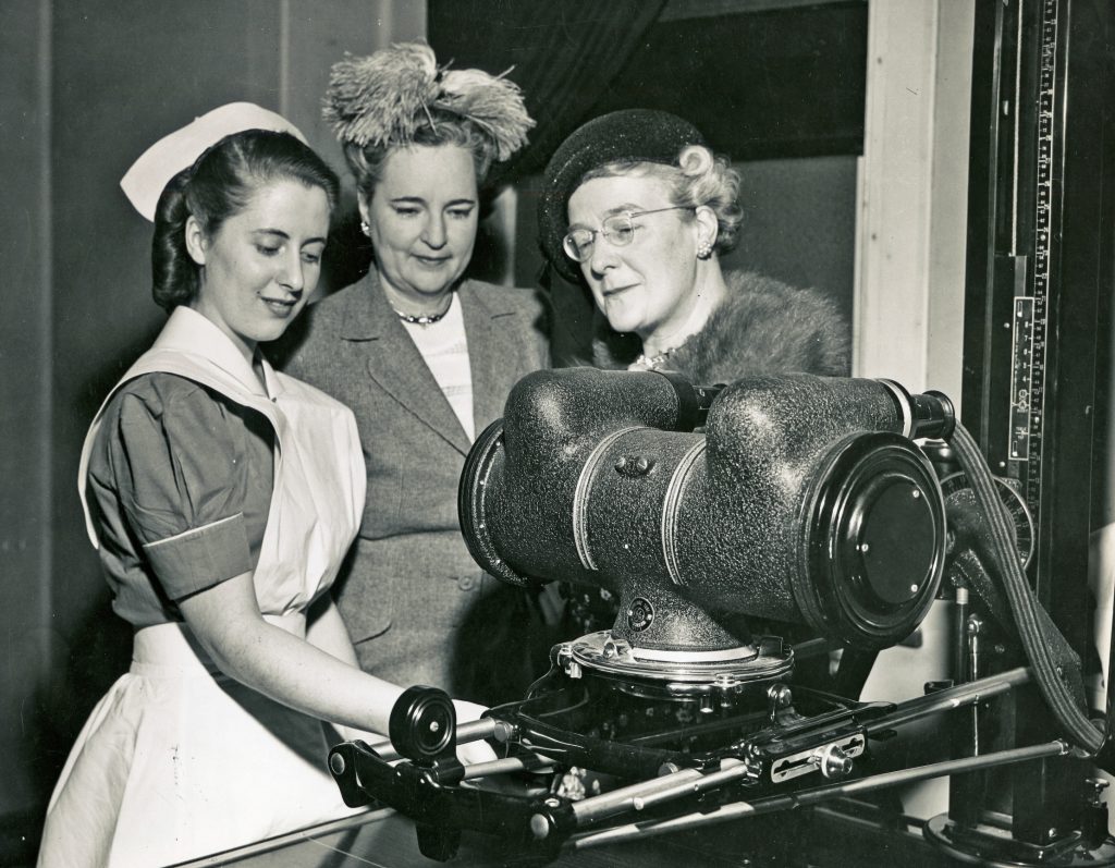 Photo en noir et blanc de trois femmes devant des appareils scientifiques. Vêtue d’un uniforme d’infirmière, la plus jeune semble démontrer l’appareil à deux femmes plus âgées élégamment vêtues.