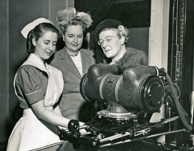 A black and white photo of three women before scientific equipment. One, who is younger and in nurses' uniform, appears to be demonstrating the machine to two elegantly dressed older women.