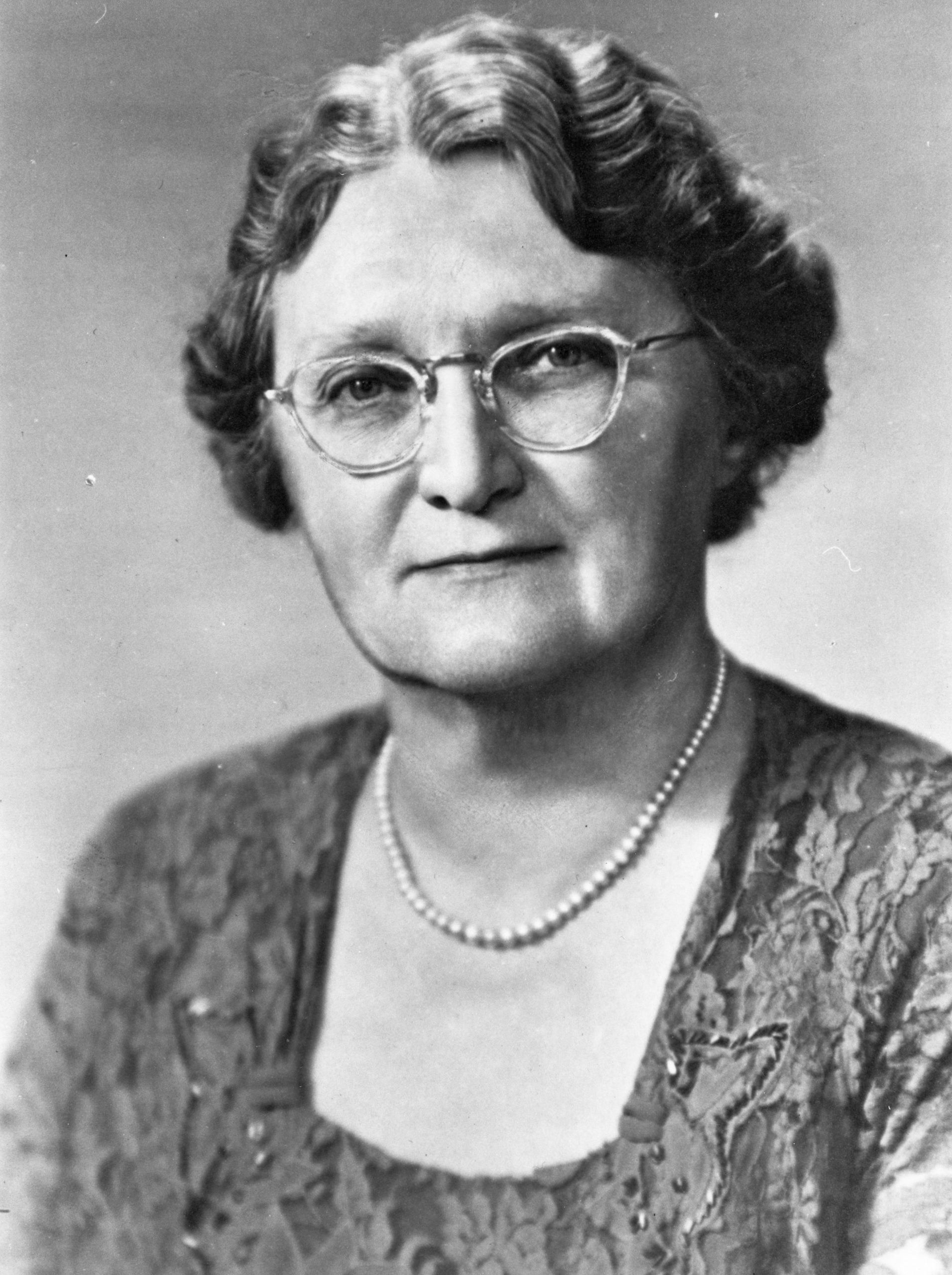 A black and white portrait of an elegantly dressed woman, wearing glasses.