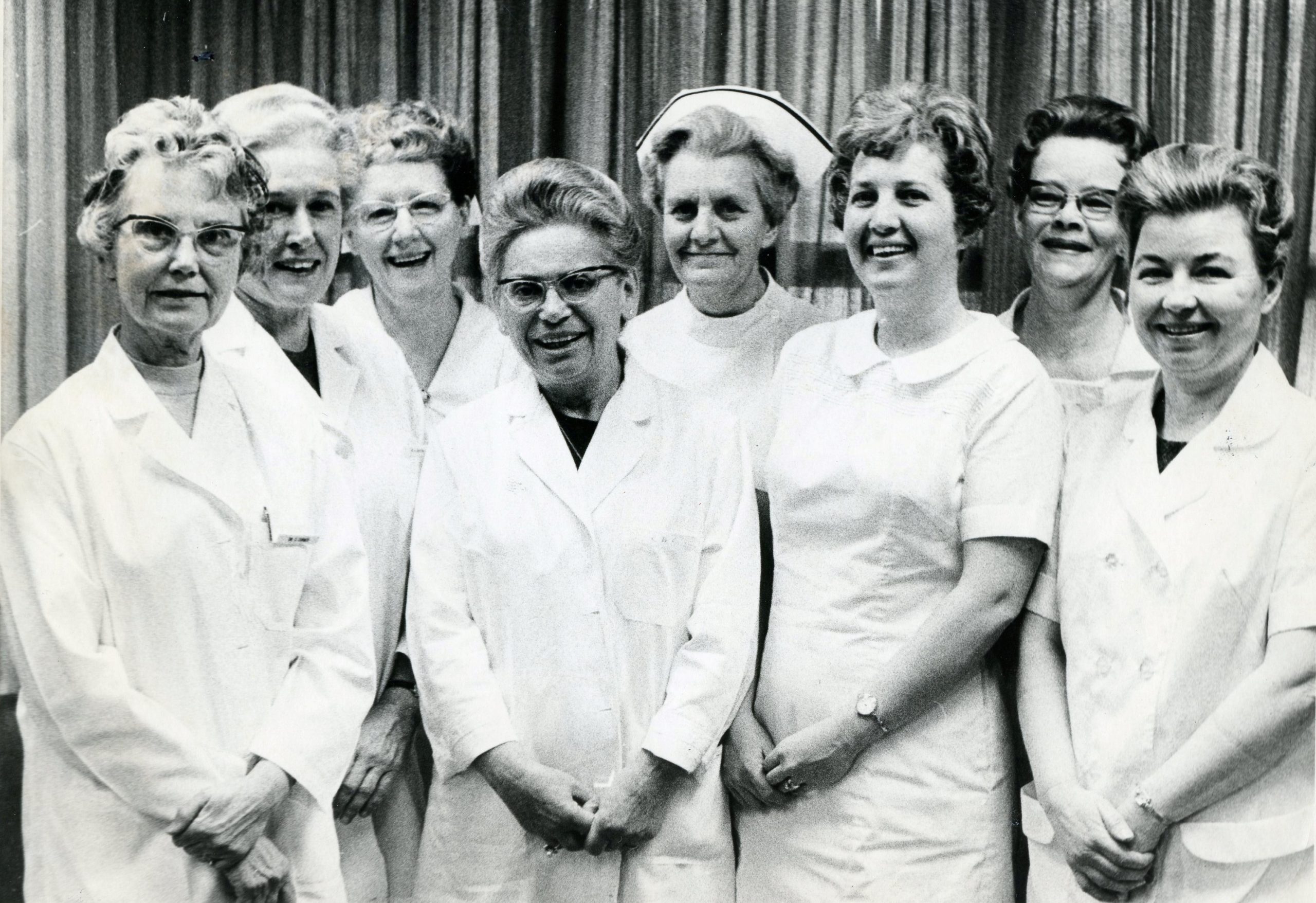 A black and white photo of eight older women in lab-coats smiling and facing the camera.