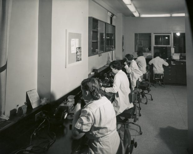 A black and white photo of a row of women in lab-coats sitting a long table and looking through microscopes.