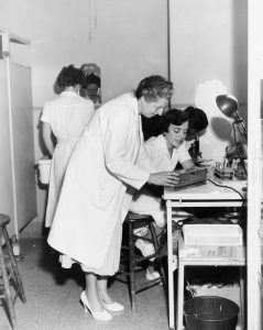 A black and white photo of women in lab-coats engaged in a variety of tasks. An older woman looks on as a younger woman works on a machine. Another woman appears to be washing up at a sink.