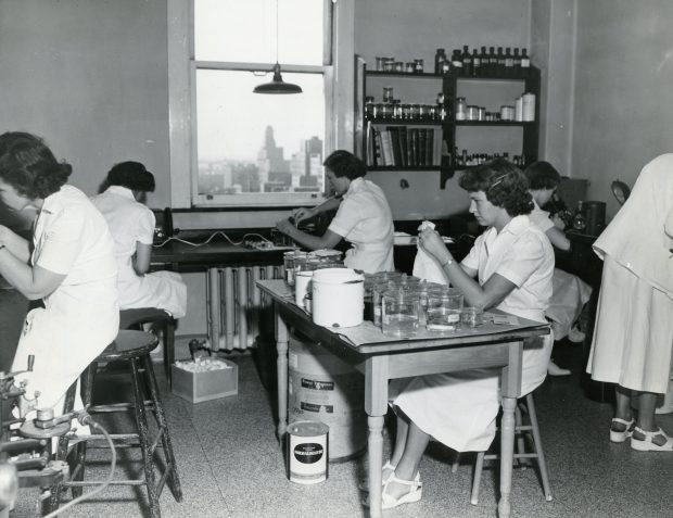 A black and white photo of young women in lab-coats work at various tasks in a laboratory.