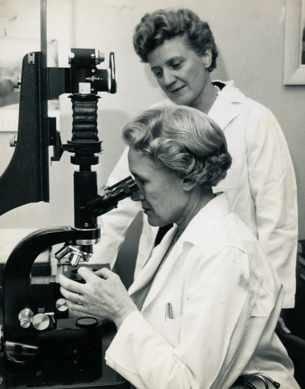 A black and white photo of two women in lab-coats. One is looking through a large microscope. The other looks on.