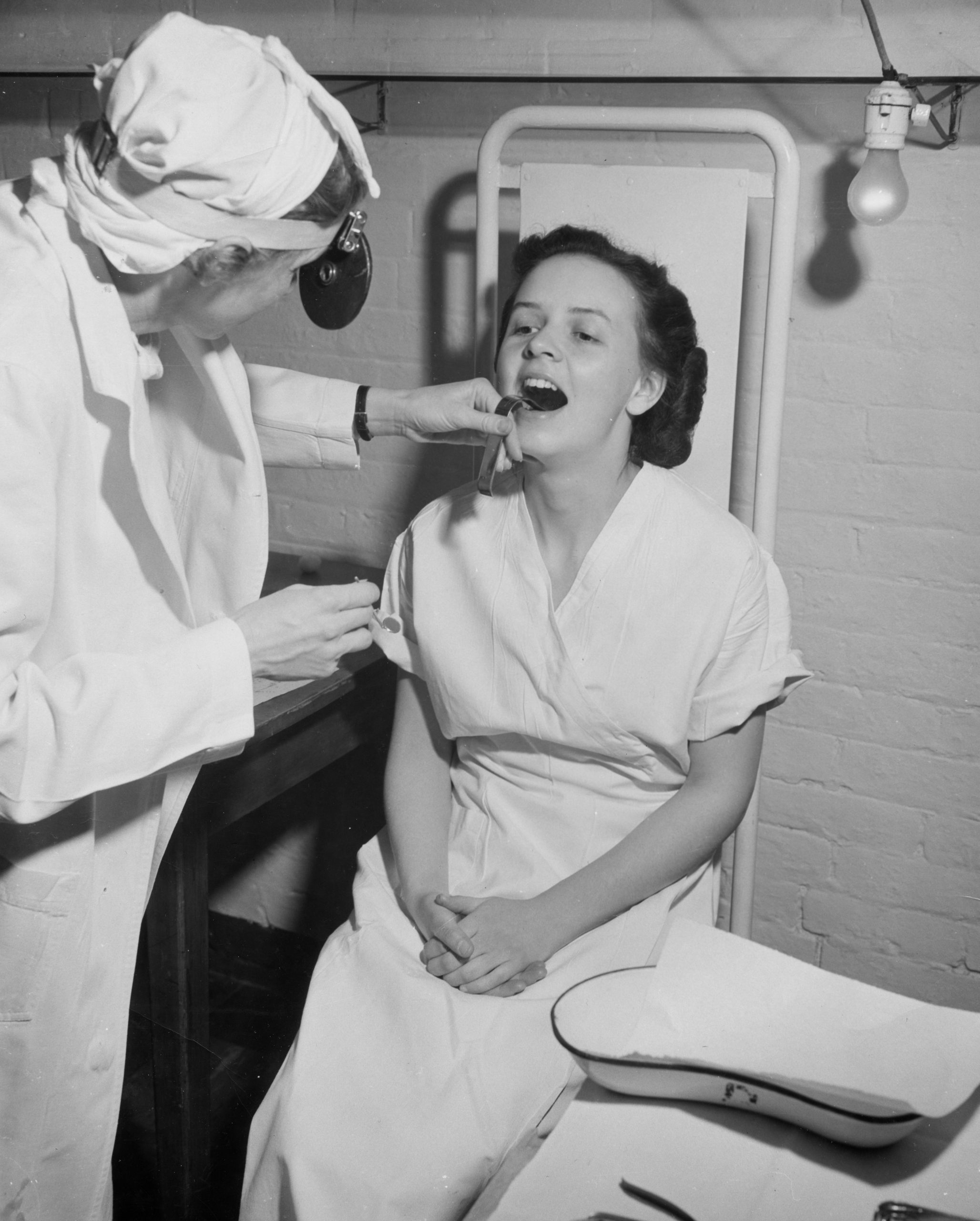 A black and white photo of a woman in scrubs with a head mirror examines the open mouth of a young woman in a hospital gown.