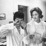 A black and white photo of two women in lab coats studying a vial that one is holding.