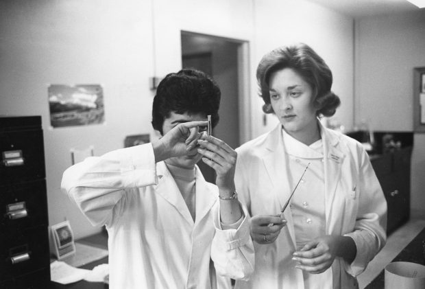 A black and white photo of two women in lab coats studying a vial that one is holding.