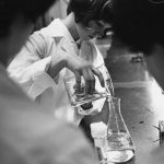 A black and white photo of a woman in a lab coat pouring a clear liquid from one beaker to another. She is framed by two other women in the foreground.