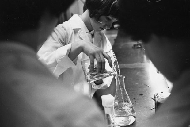 A black and white photo of a woman in a lab coat pouring a clear liquid from one beaker to another. She is framed by two other women in the foreground.